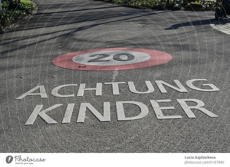 Straße mit Schild für Geschwindigkeitsbegrenzung 20 Kilometer pro Stunde und Aufschrift in deutscher Sprache: Achtung Kinder. Die Geschwindigkeitsbegrenzung und die Aufschrift sind auf dem Boden markiert.