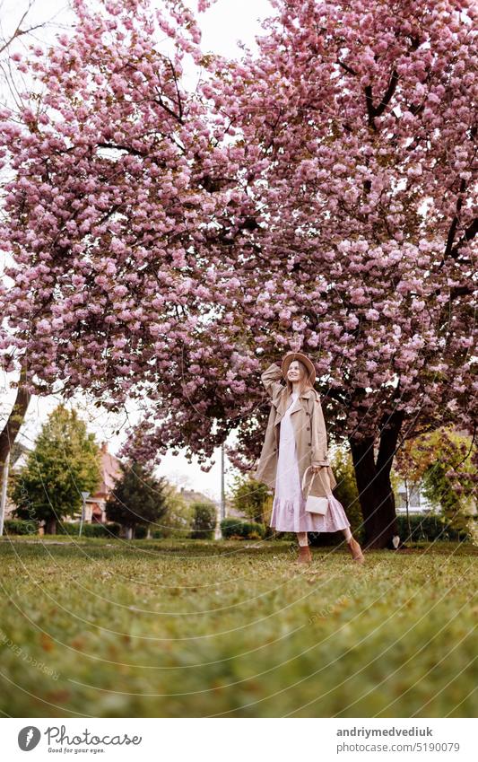 Tag der Frau. Schöne Frau in der Nähe der Sakura-Bäume. Frau in Hut, Kleid und stilvollem Mantel. Rosa Blumen blühen in Uzhhorod, Ukraine. Blüte um. Frühlingszeit Konzept.