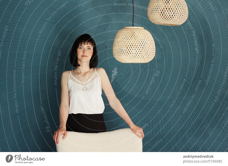 Millennial junge brünette Frau schaut in die Kamera und steht in kreativen Büro mit Grange blau-grüne Wand, weichen Stuhl und Rattan-Lampen. Erfolgreiche selbstbewusste Designerin. copy space