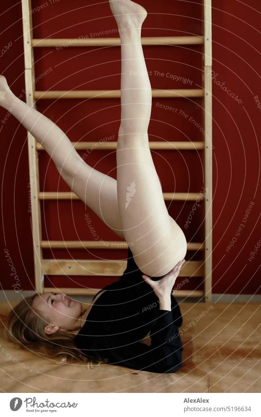 eine junge, blonde, schlanke Frau in schwarzem Body macht Gymnastik auf einer Turnmatte vor einer roten Wand mit Sprossenwand sportlich schön lächeln fit