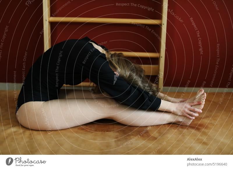 eine junge, blonde, schlanke Frau in schwarzem Body macht Gymnastik auf einer Turnmatte vor einer roten Wand mit Sprossenwand sportlich schön lächeln fit