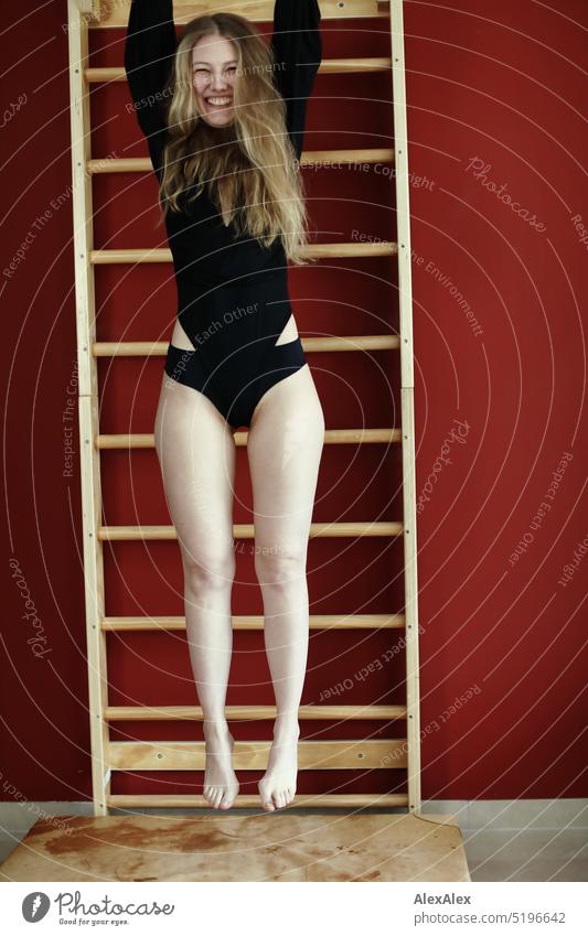eine junge, blonde, schlanke Frau in schwarzem Body hängt an der Sprossenwand über einer Turnmatte vor einer roten Wand sportlich schön lächeln fit