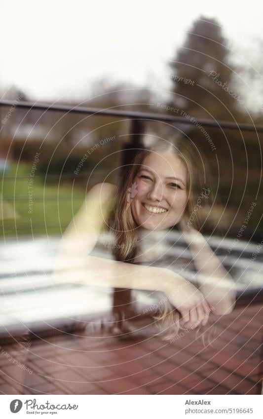 eine junge, blonde, schlanke Frau in schwarzem Kleid liegt hinter der Balkonfensterscheibe auf der Couch und lächelt in die Kamera sportlich schön lächeln fit