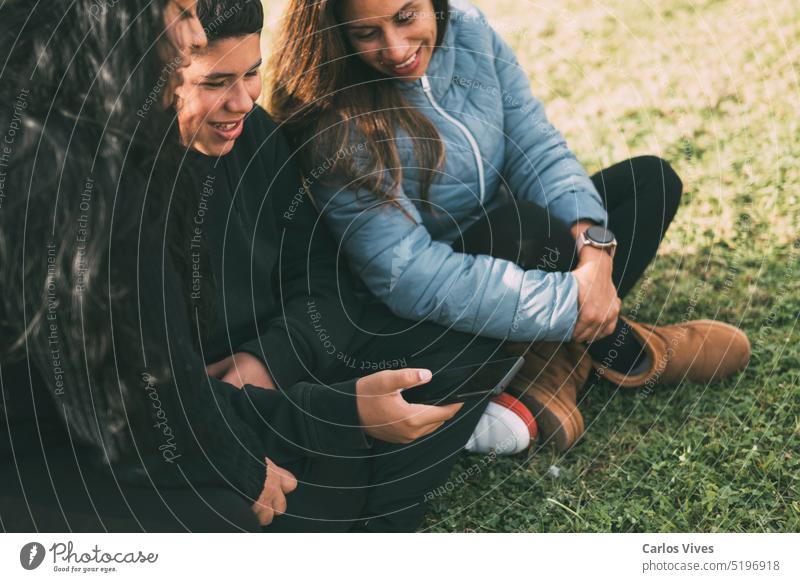 Hispanischer männlicher Teenager, der sein Smartphone in der Hand hält und wegschaut, während er mit seiner hispanischen Mutter und seiner Schwester an einem sonnigen Tag im Park im Gras sitzt