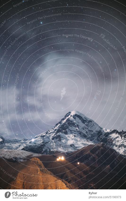 Stepantsminda, Georgien. Night Starry Sky With Glowing Stars Above Peak Of Mount Kazbek Bedeckt mit Schnee. Berühmte Gergeti Kirche in der Nacht Blitz. Schöne georgische Landschaft im Spätherbst