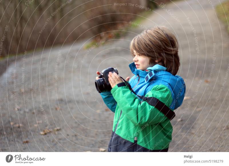 PC-Nachwuchs - Porträt eines Jungen, der eine Kamera in der Hand hält und lächelnd aufs Display schaut Mensch Kind fotografieren schauen stehen knipsen Jacke