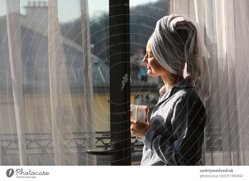 Schöne junge Frau mit Badetuch auf dem Kopf hält eine weiße Tasse mit heißem Kaffee oder Tee zu Hause am Fenster. Lächelndes Mädchen nach der Dusche genießt Aroma Getränke und haben Frühstück am Morgen