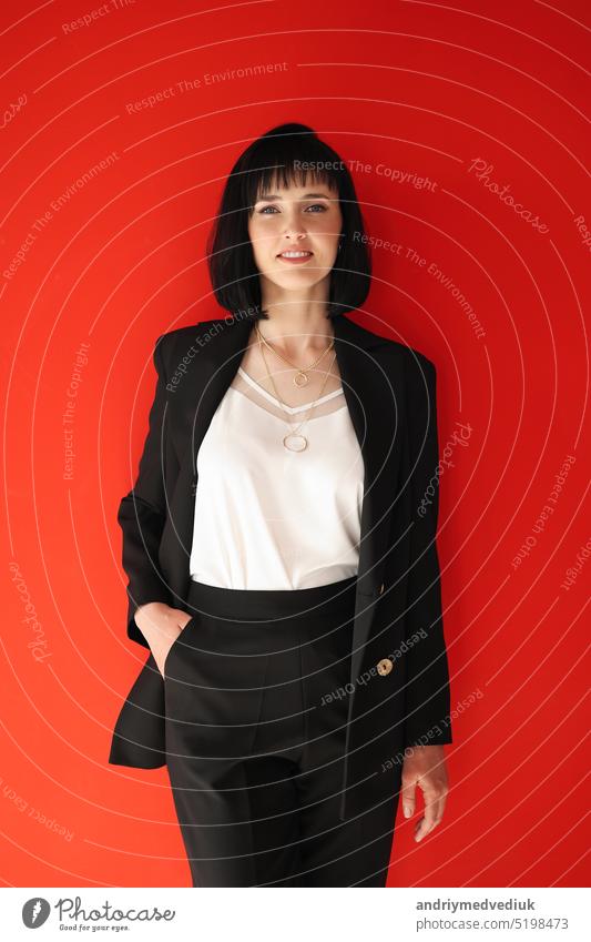 Schöne weibliche brünette Geschäftsfrau CEO in einem schwarzen Anzug, isoliert auf rotem Hintergrund, selbstbewusst stehend mit Armen in den Taschen. Frauen in der Wirtschaft.