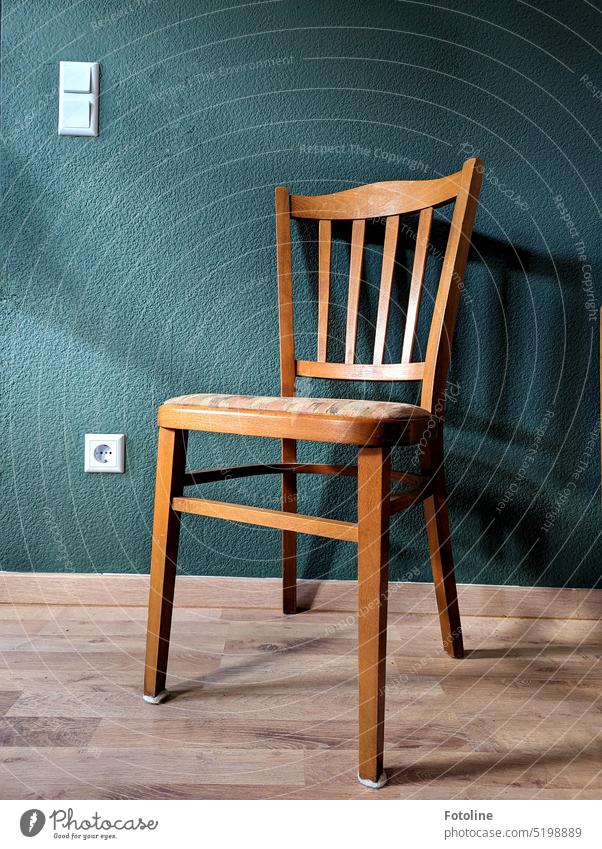 Der leere Stuhl vor einer petrolfarbenen Wand auf Laminatboden wirft seinen Schatten an die Wand. Steckdose und Lichtschalter leuchten weiß. Petrolfarben Holz