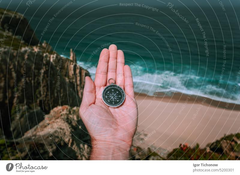 Blick auf den Kompass in der Hand eines Mannes, der das Konzept der Abenteuersuche gegen das Meer und die Wellen der Küstenlinie im Hintergrund symbolisiert