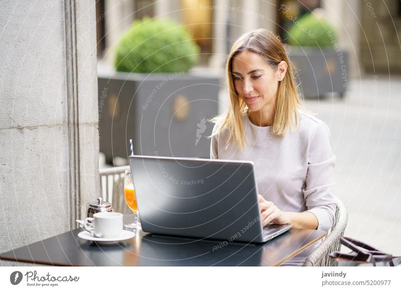 Lächelnde selbständige Frau mit Laptop in einem Cafe Arbeit Café freiberuflich Tippen abgelegen positiv Straße Terrasse Kaffee Erwachsener blond benutzend
