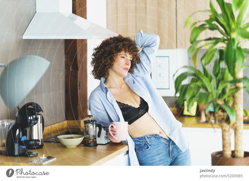 Heiße Frau in schwarzem Dessous-Shirt und Jeans mit Tasse Tee Haare berühren Tastkopf Teetrinken Heißgetränk aufknöpfen Küche Erwachsener krause Haare Freizeit