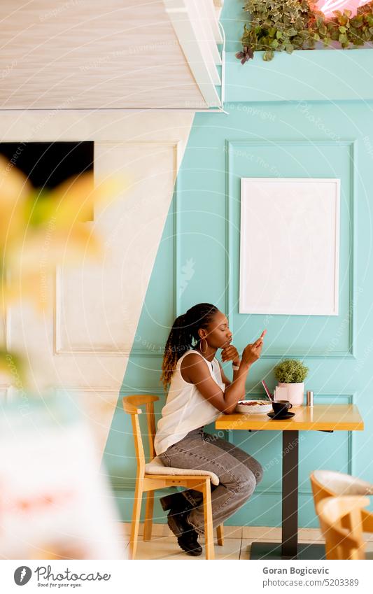 Junge schwarze Frau, die ein Mobiltelefon benutzt, während sie in einem Café frühstückt Person jung Mobile Kaffee Dame Afro-Look schön lässig Telefon Frühstück