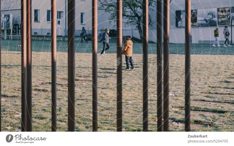 Gitterstreben teilen das Bild vertikal, dahinter eine Wiese in der Stadt, spielende Menschen Streben städtisch Mauerpark Park laufen Sonne sonnig Leben Freizeit