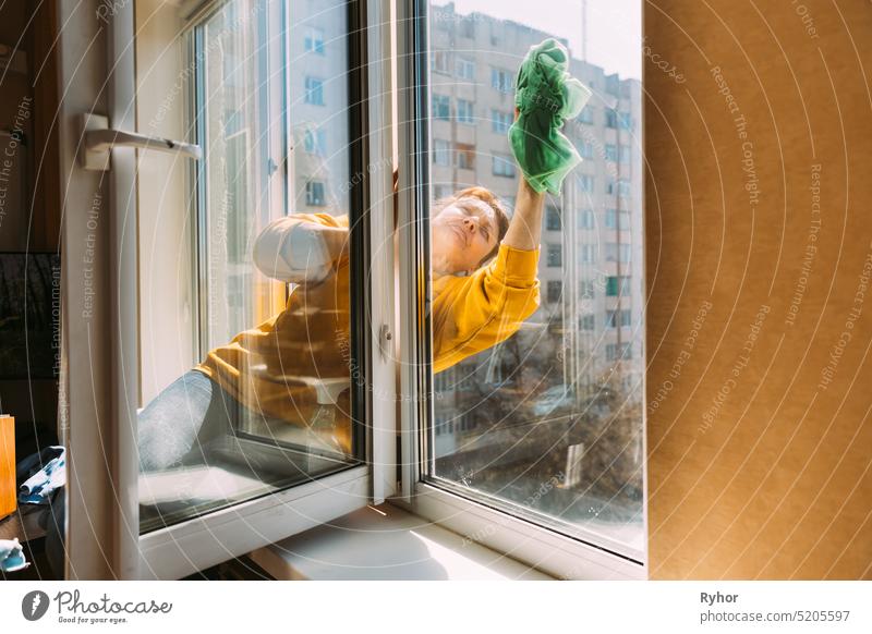 Kaukasische Frau von fünfzig in einem gelben Pullover und Jeans wäscht ein staubiges Fenster in der Wohnung. Eine 50 Jahre alte Frau reinigt Fenster von Flecken mit Lappen. Ältere Frau reinigt Haus, macht Hausarbeit