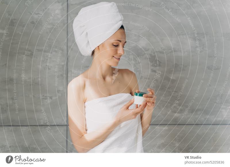 Junges Mädchen im Handtuch hält Kosmetiktopf mit Creme im Badezimmer. Skincare Schönheitsprodukte Werbung Frau Hautpflege Sahne Serum Glas Behandlung natürlich