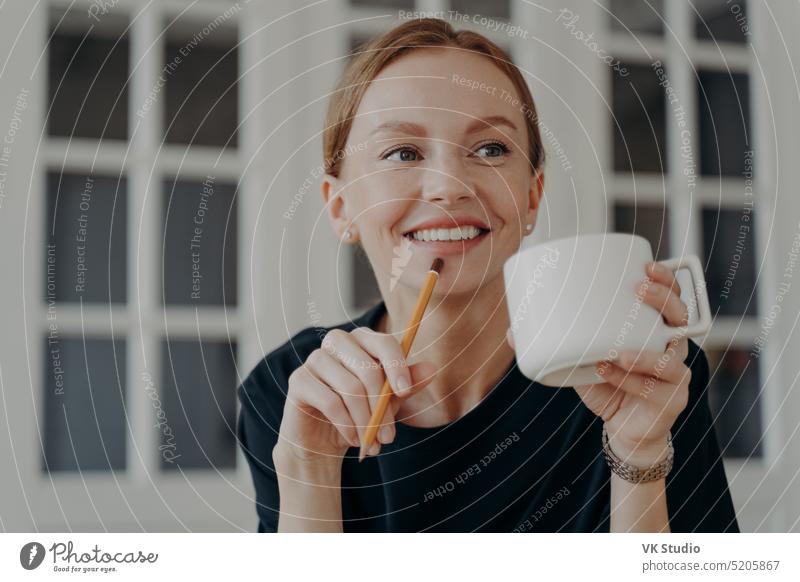 Eine erfolgreiche Geschäftsfrau mittleren Alters trinkt Tee, lächelt und träumt am Arbeitsplatz. Frau Kaffee träumend Tasse trinken Bleistift Hand Porträt