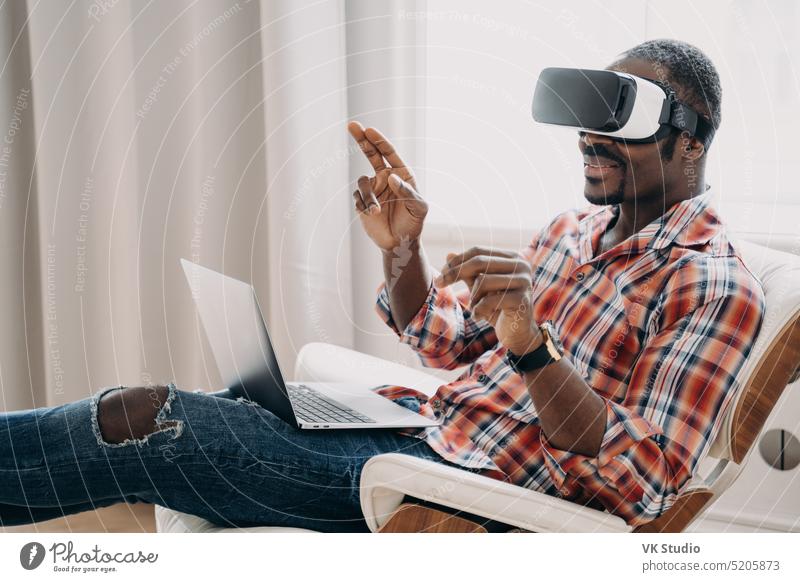 Afroamerikanischer Mann mit Virtual-Reality-Brille arbeitet oder spielt ein Videospiel auf einem Laptop, sitzt auf einem Stuhl Afroamerikaner virtuell Realität