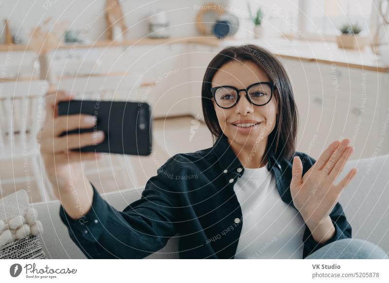 Eine Frau mit Brille nimmt einen Videoanruf entgegen, hält ein Smartphone in der Hand und grüßt mit winkender Hand, sitzt zu Hause freundlich Lifestyle