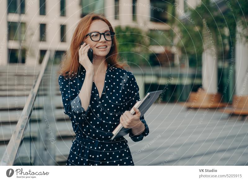 Positive rothaarige europäische Frau führt ein Telefongespräch beim Gehen im Freien Business Person Büro Gesicht Technik & Technologie jung laufen sprechend