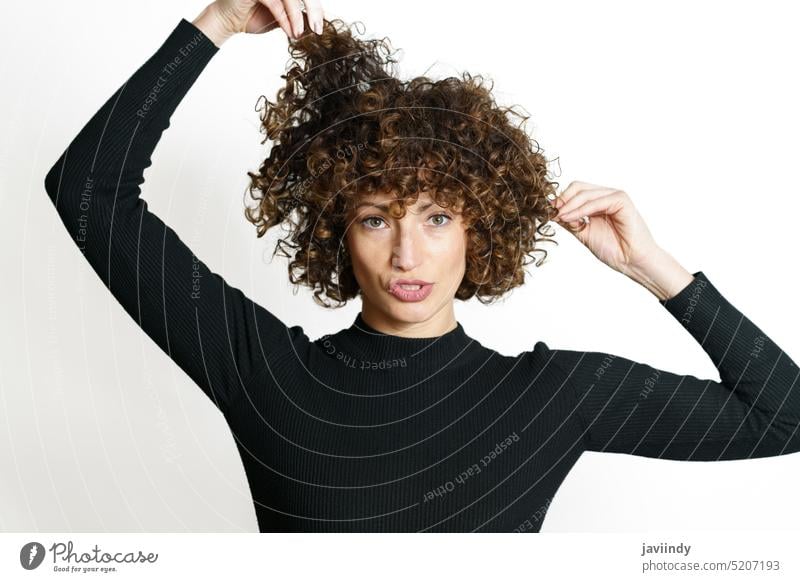 Junge Frau berührt lockiges Haar vor weißer Wand Model ziehen krause Haare Mund geöffnet Pullover Haare berühren expressiv attraktiv Windstille Stil Vorschein