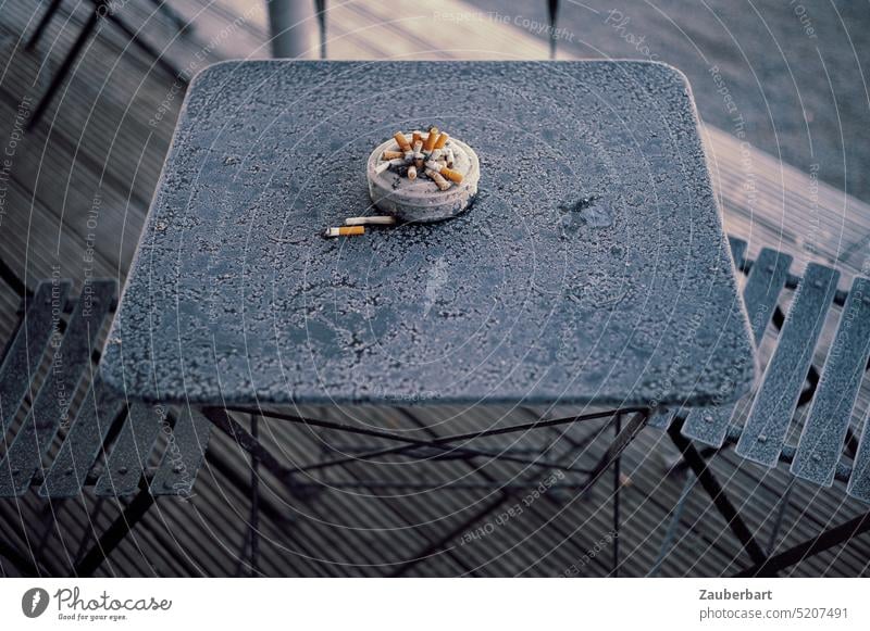 Voller Aschenbecher mit Zigarettenstummeln auf eisigem Gartentisch rauchen Stummel aufhören stop Tisch kalt draußen rausgehen Vorsatz gut ungesund Nikotin