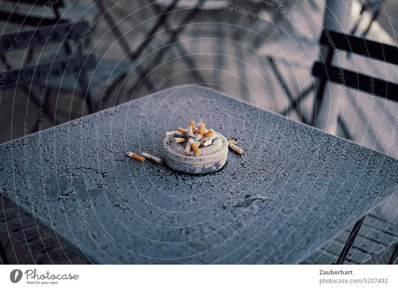 Voller Aschenbecher mit Zigarettenstummeln auf eisigem Gartentisch rauchen Stummel aufhören stop Tisch kalt draußen rausgehen Vorsatz gut ungesund Nikotin