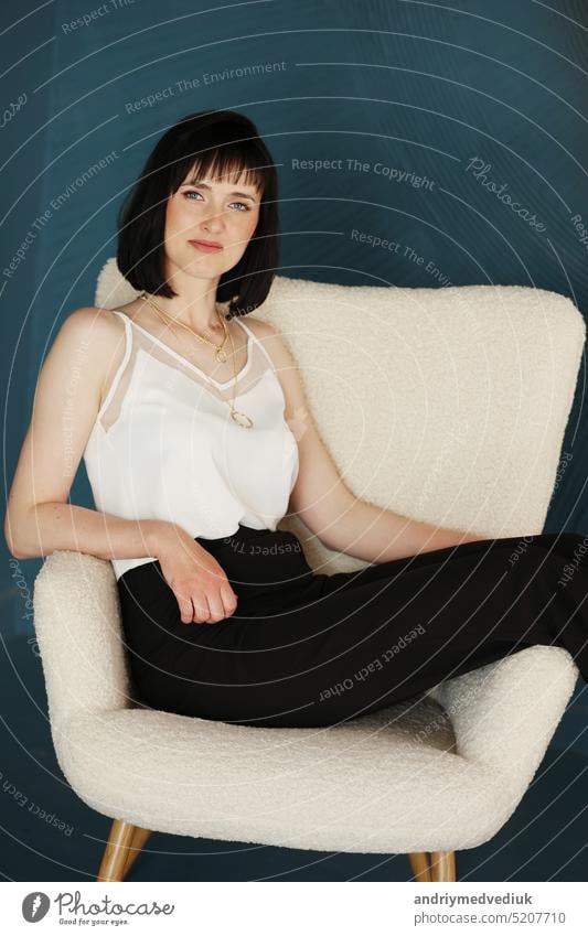 attraktive Business-Frau sitzt in bequemen weißen Stuhl. Lächelnde brünette Dame mit kurzen Haaren trägt schwarze Büro Hose und weiße Bluse, High-Heels isoliert auf blau-grünem Hintergrund. copy space.