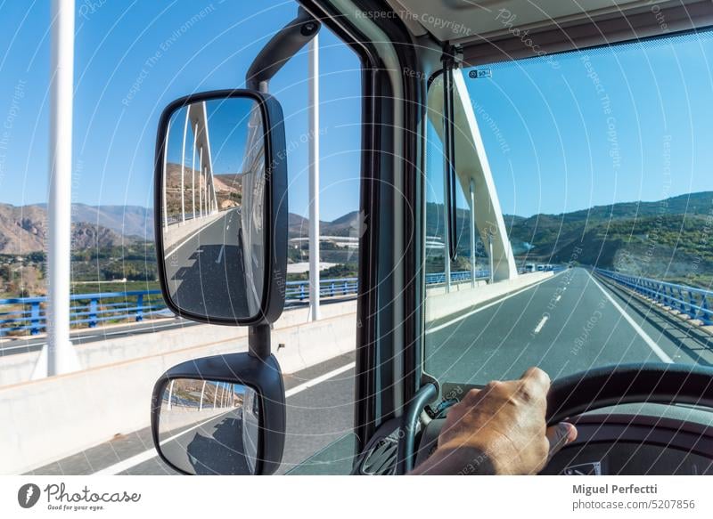 Blick aus der Fahrerposition eines Lkw auf die Straße, auf der er fährt, wenn er über eine Brücke fährt, und auf die Rückspiegel, in denen sich die Brücke spiegelt.