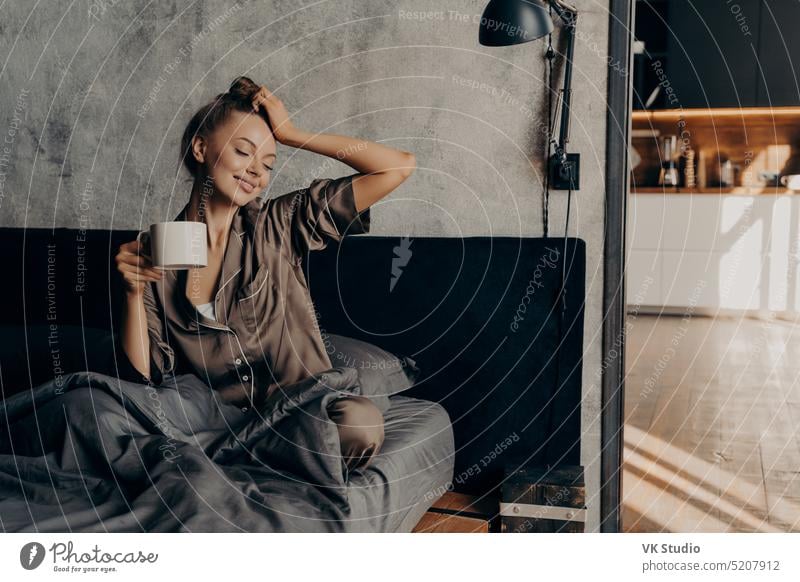 Porträt einer jungen entspannten Frau in stilvollem Hausanzug, die mit einer Tasse Kaffee im Bett aufwacht stylisch heimwärts Anzug Aufwachen nach oben