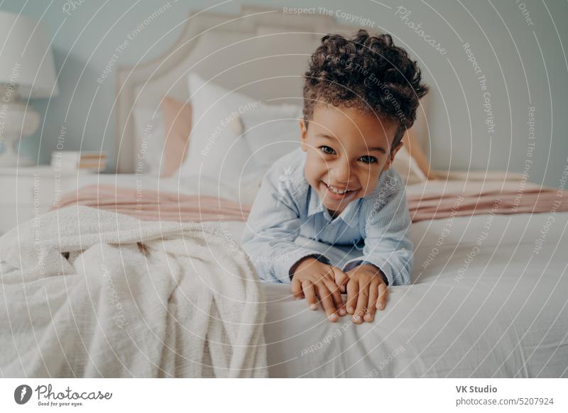 Glücklicher lockiger afroamerikanischer Junge liegt auf dem Bett und lächelt in die Kamera niedlich wenig Kind gemischt Rennen Afrikanisch Amerikaner Porträt