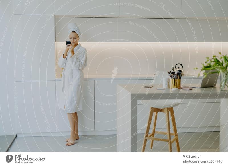 Full length shot of young beautiful woman dressed in white bathrobe applies face cream looks in mirror takes care of her skin poses near kitchen furniture stands bare feet. Menschen Schönheit Gesichtspflege