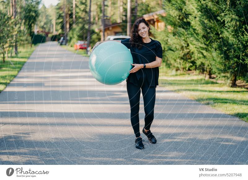 Fitness Frau Übungen mit Fitness-Ball im Freien Spaziergänge auf der Straße in der Nähe von Wald hat einen gesunden Lebensstil in schwarzer aktiver Kleidung gekleidet, hat glücklichen Ausdruck, bleibt in guter körperlicher Verfassung. Fitness-Konzept
