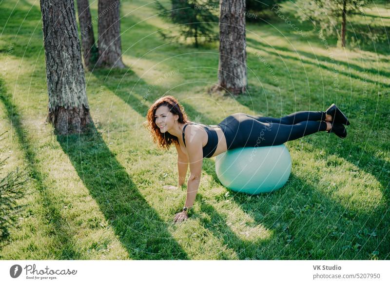 Outdoor-Aufnahme von aktiven Brünette Frau Übungen mit Fitness-Ball posiert auf grünem Gras, in aktive Kleidung gekleidet, genießt sonnigen Tag und frische Luft im Park, hält fit, macht Gymnastik-Übungen.