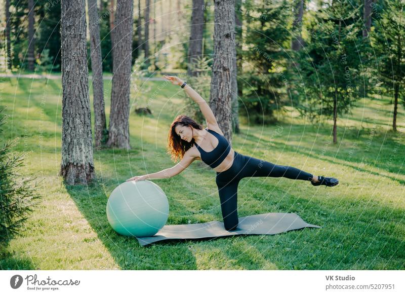 Sport und aktiver Lebensstil Konzept. Schlanke sportliche Frau in schwarzer Kleidung lehnt sich zur Seite auf Fitness-Ball, hebt die Arme, posiert auf Karemat im grünen Park, trainiert Yoga im Freien, hat athletische Körperform.