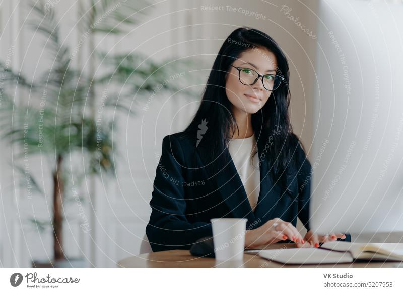 Seriöse erfolgreiche weibliche Büroangestellte in formellem Outfit, trägt eine Brille, beschäftigt mit Online-Arbeiten, posiert am Schreibtisch, arbeitet am Computer, liest Business-News oder schaut Tutorial, genießt den Arbeitsprozess.