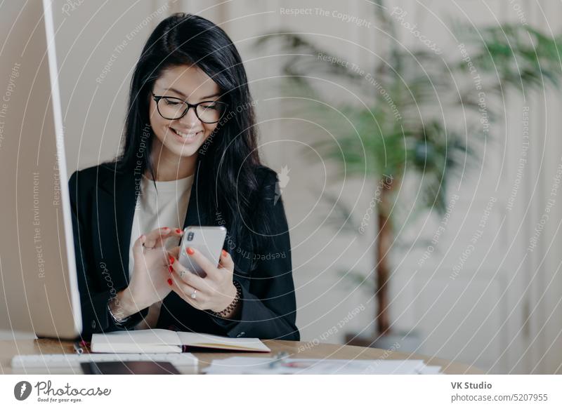 Hübsche lächelnde Freiberuflerin arbeitet aus der Ferne, konzentriert sich auf ihr Smartphone, sendet Feedback auf empfangene Nachrichten, sitzt in einem Coworking Space, formell gekleidet, arbeitet am Computer, surft im Internet