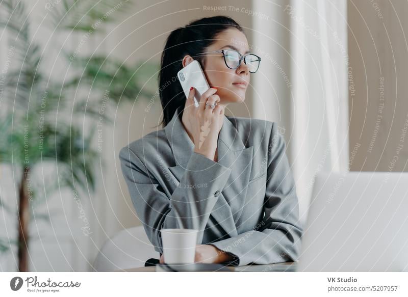 Foto einer Werbetexterin führt ein Telefongespräch, schaut zur Seite, plaudert über Ideen für eine neue Publikation, förmlich gekleidet, posiert vor einem Laptop im Coworking Space, diskutiert den Arbeitsprozess