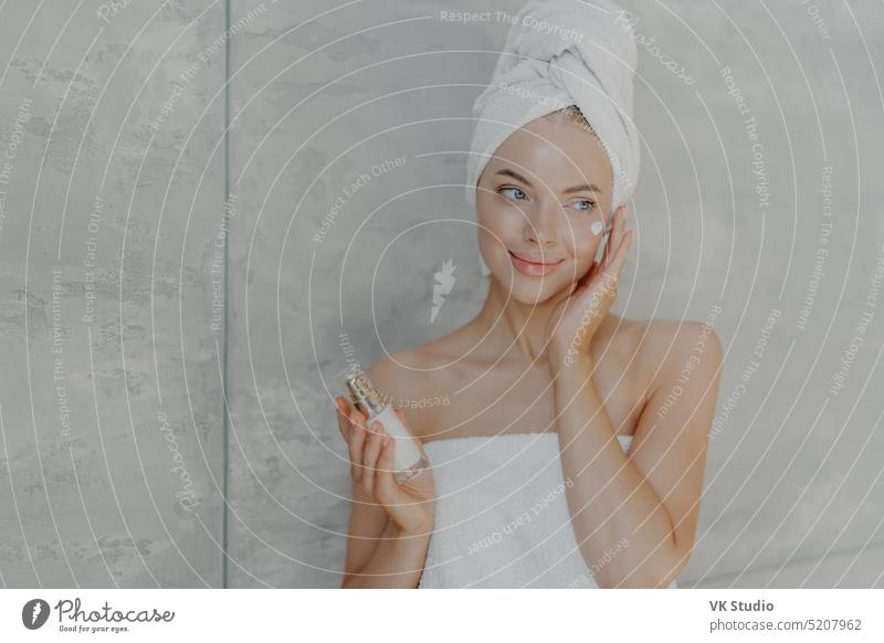 Lovely junge Frau gilt Schönheit Lotion auf Gesicht hat zufriedenen Ausdruck trägt Badetuch auf dem Kopf posiert mit nackten Schultern isoliert über grauen Hintergrund. Kosmetologie Wohlbefinden und Spa-Konzept