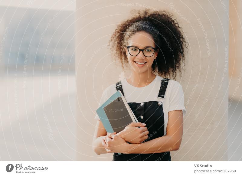 Eine hübsche, lockige Studentin trägt ein weißes Freizeithemd und eine Latzhose, hält einen Notizblock oder ein Lehrbuch in der Hand und blickt durch eine transparente Brille auf einen weißen Hintergrund. Menschen und Lebensstil