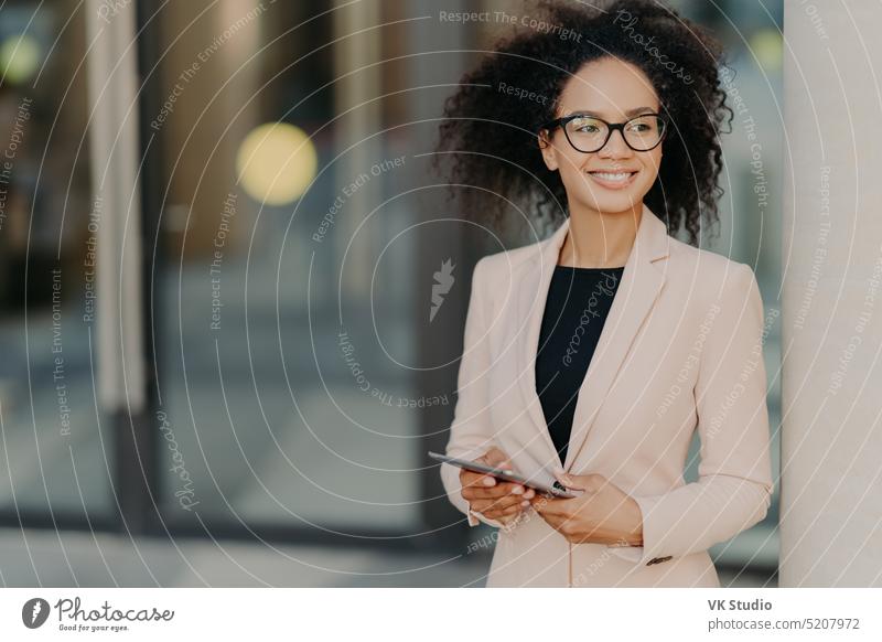 Positive erfolgreiche Unternehmerin mit Afro-Haar hält digitales Tablet, steht im Freien in der Nähe des Bürogebäudes, trägt formelle Kleidung, schaut weg, wartet in der Pause auf einen Kollegen, der zu Abend isst