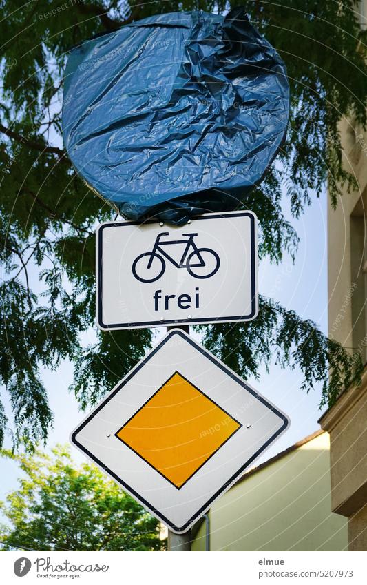 Verkehrszeichen Vorfahrtstraße, Zusatzzeichen Radverkehr frei und ein mit blauer Plastikplane verhülltes rundes Verkehrszeichen an einem Metallrohr unter einem Baum und neben einem Wohnhaus