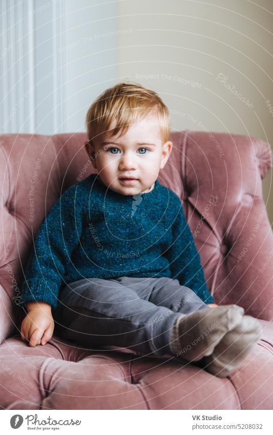 Vertikales Porträt eines unschuldigen kleinen Kindes mit blauen Augen und prallen Wangen, schaut direkt in die Kamera, wartet auf Kinder, die Geburtstag haben, sitzt in einem bequemen rosa Sessel im Wohnzimmer. Kinder
