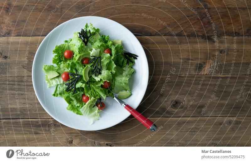 Weißer Teller mit gemischtem Salat mit Algen, Tomaten, Kopfsalat und Avocado. Hintergrund aus Holz. Platz zum Kopieren. Holzboden Gabel Besteck weiße Platte