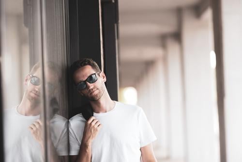 Der junge Mann mit Sonnenbrille lehnt lässig an einer Wand und schaut nachdenklich in die Kamera Junger Mann Maskulinität Lässigkeit Mannlich Gesicht Denken