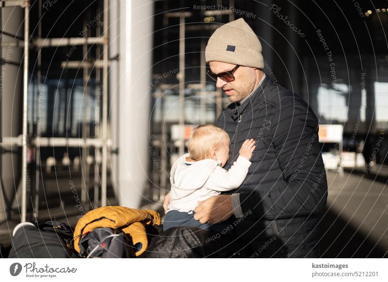 Fatherat tröstet seine weinenden Säugling Junge Kind müde sitzen auf der Oberseite des Gepäcks Wagen vor dem Flughafen-Terminal Station während der Reise wih Familie.