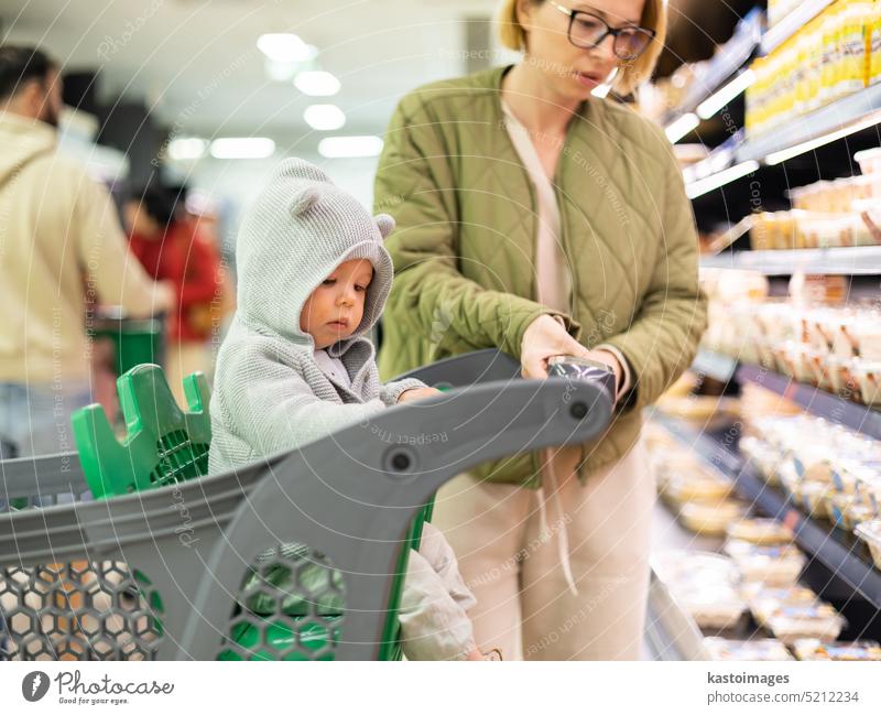 Casualy gekleidet Mutter Auswahl von Produkten in der Abteilung des Supermarktes Lebensmittelgeschäft mit ihrem Säugling Junge Kind im Einkaufswagen. Frau jung