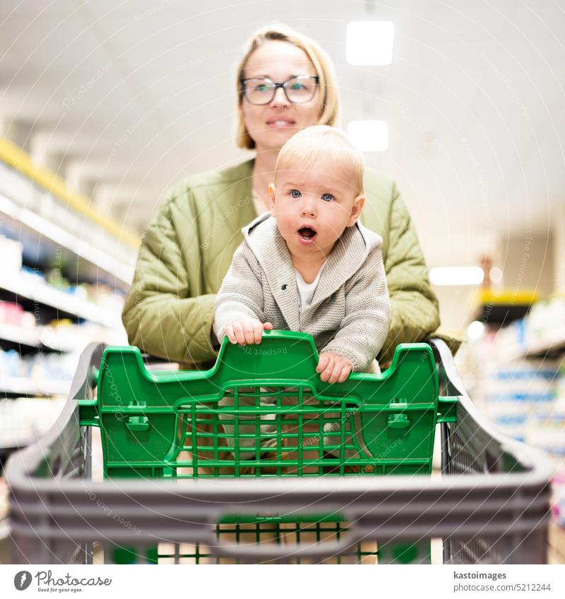 Mutter schiebt Einkaufswagen mit ihrem Säugling Baby Junge Kind nach unten Abteilung Gang im Supermarkt Lebensmittelladen. Einkaufen mit Kindern Konzept. Frau