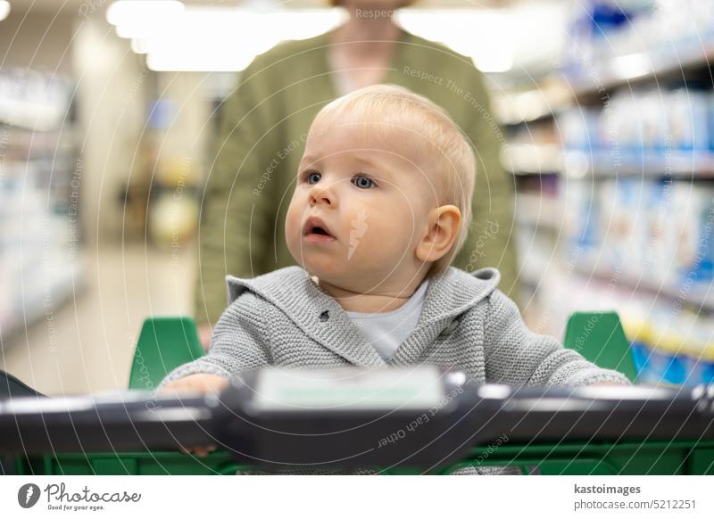Mutter schiebt Einkaufswagen mit ihrem Säugling Baby Junge Kind nach unten Abteilung Gang im Supermarkt Lebensmittelladen. Einkaufen mit Kindern Konzept. Frau