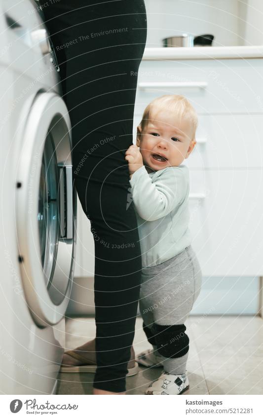 Kleines Baby Junge Kind versteckt zwischen Mütter Beine fordern ihre Aufmerksamkeit, während sie multitasking ist, versuchen, einige Hausarbeit in der Küche zu Hause zu tun. Mutter im Mutterschaftsurlaub.